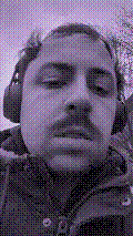 a selfie of a white person, wearing either headphones or a helmet, making an odd expression. the photo is edited to be colorized into a monochrome purple-ish color palette.